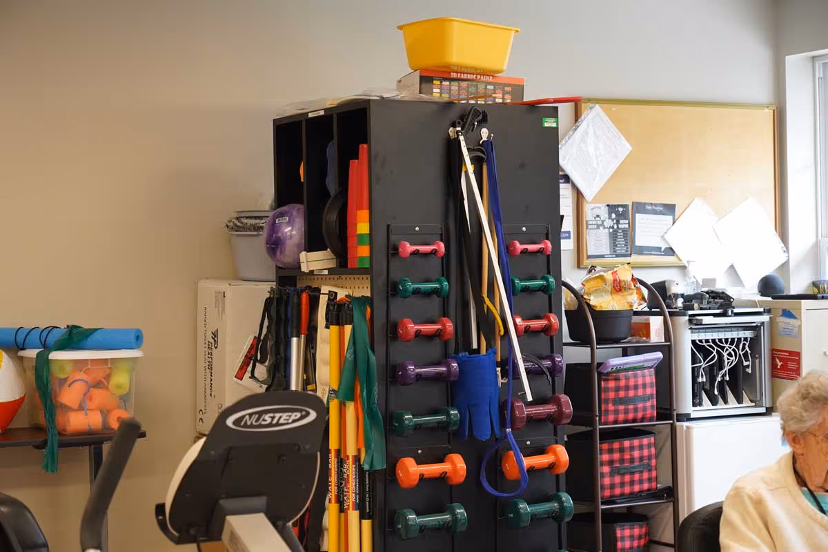 A room with various exercise equipment including colorful dumbbells, resistance bands, and other fitness tools organized on a black storage rack. A person with gray hair is partially visible sitting in the lower right corner. There is a bulletin board with papers pinned on the wall and a window letting in natural light.