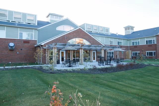 Exterior view of a senior living facility building with a brick and siding facade, featuring a covered patio area with tables, chairs, and umbrellas. The building has multiple windows and a well-maintained lawn with some small plants and shrubs in the foreground.