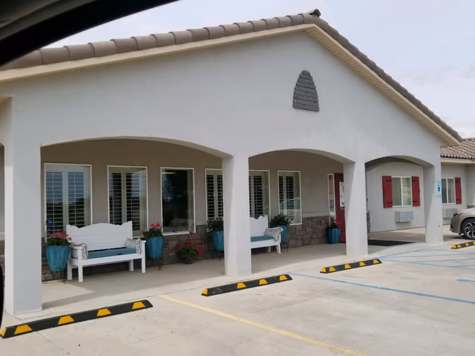 Exterior view of a single-story building with white walls and a tiled roof. The building has a covered porch with two white benches, blue flower pots with plants, and windows with white shutters. There are parking spaces in front, including a handicapped parking spot, and a car is partially visible on the right side.