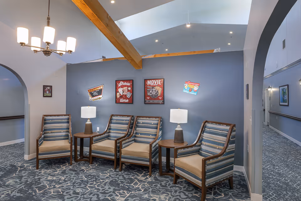 A seating area in a senior living facility with four cushioned armchairs arranged around two small round tables with lamps. The walls are painted gray and decorated with vintage-style movie posters. The ceiling features exposed wooden beams and a modern chandelier. There are arched doorways leading to hallways on either side.