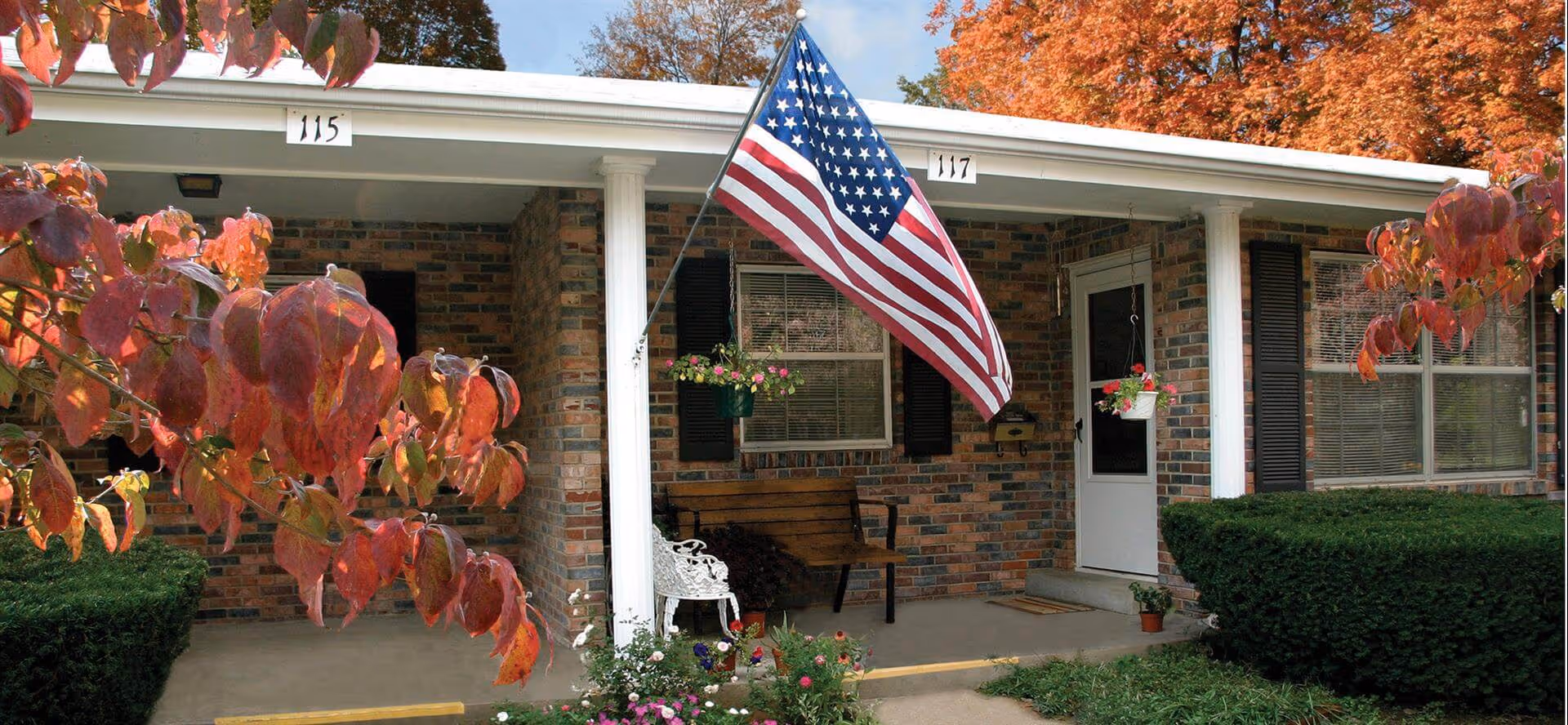 Brick front porch of a residential unit with an American flag, hanging flower baskets, a bench, and autumn foliage.
