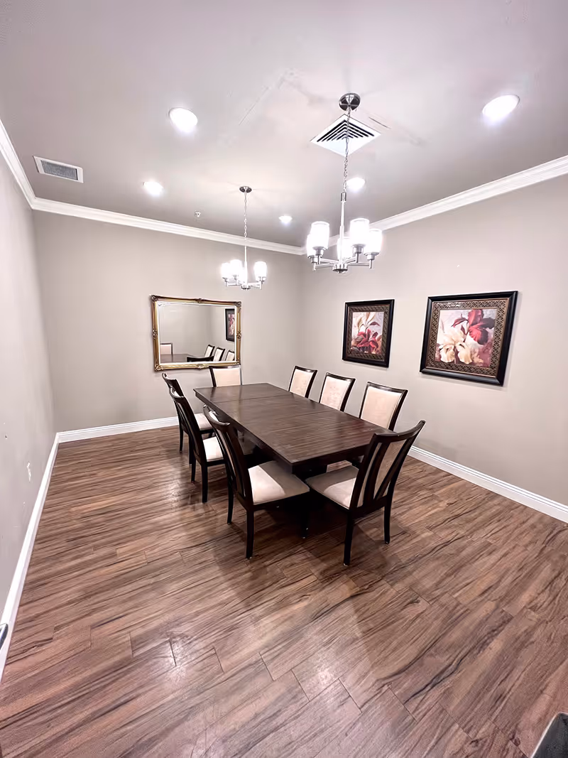 A dining room with a long rectangular wooden table surrounded by eight cushioned chairs. The room has wood flooring, beige walls, two framed floral paintings, a large wall mirror, and two modern chandeliers hanging from the ceiling.