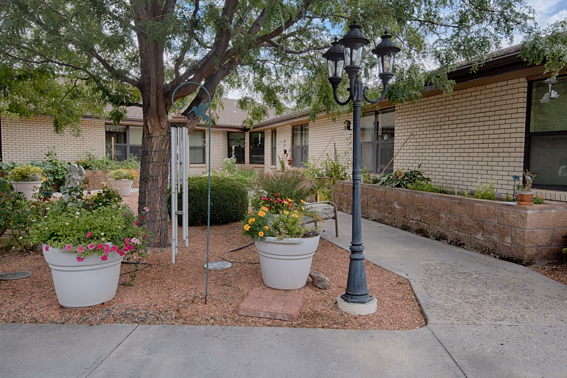 Outdoor garden area at Horizons Care Center featuring large potted flowering plants, a tree with hanging wind chimes, a black vintage-style street lamp, and a paved walkway alongside a brick building with windows.