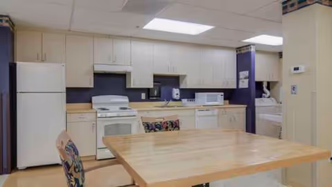 A bright communal kitchen with white cabinets, appliances, a wooden dining table, and chairs.