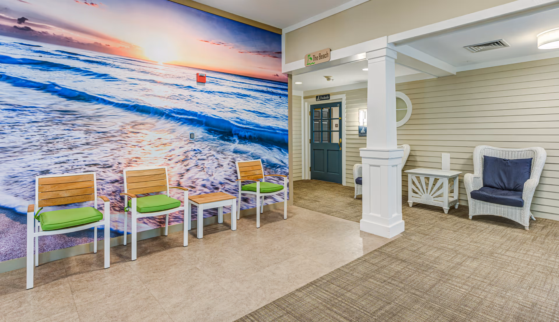 Interior seating area featuring a large beach mural, three wooden chairs with green cushions, wicker armchairs, and a blue door under a sign reading 'The Beach'.