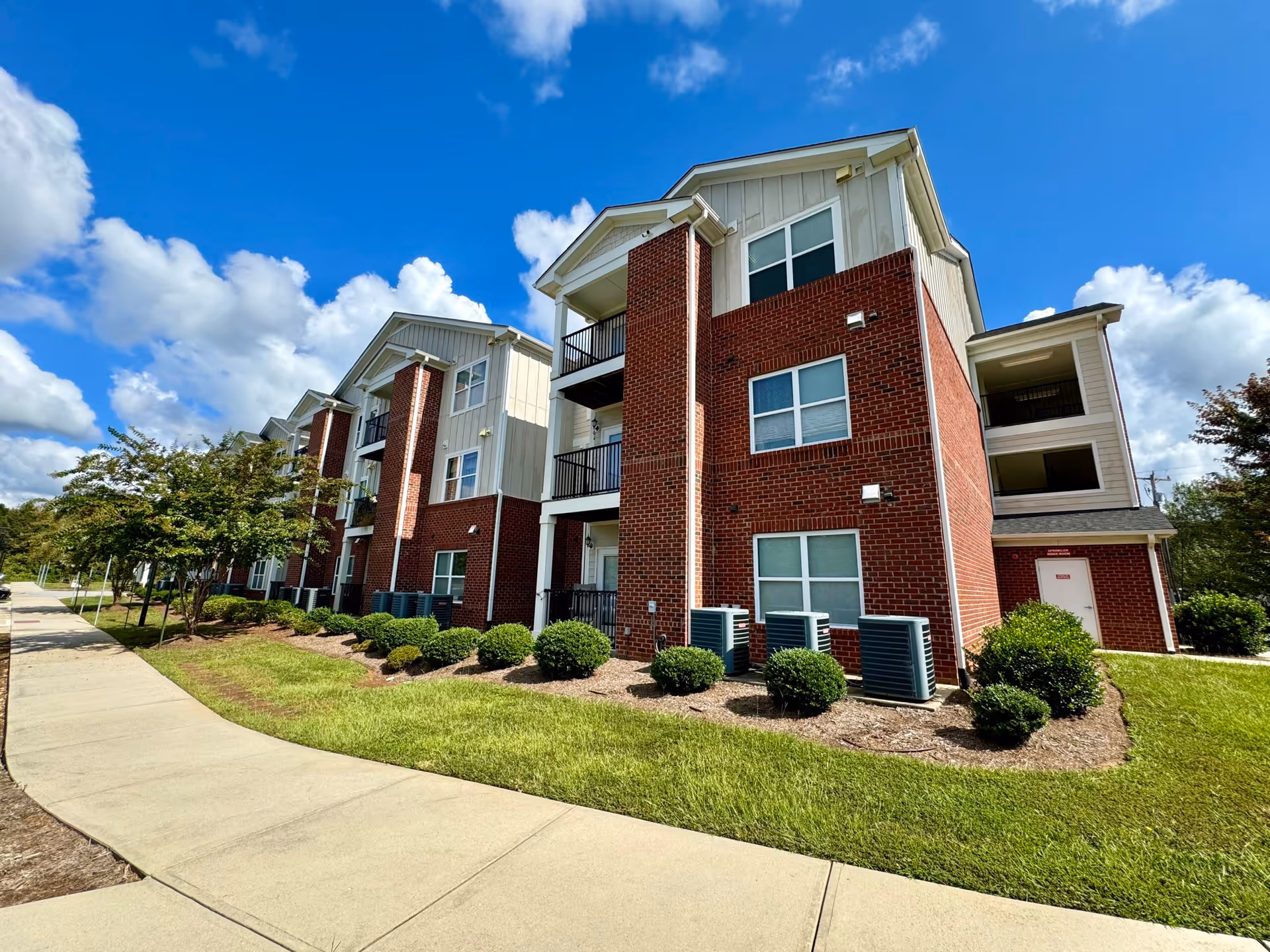 Three-story brick-and-siding apartment building with balconies, landscaped shrubs, and a sidewalk under a blue sky.
