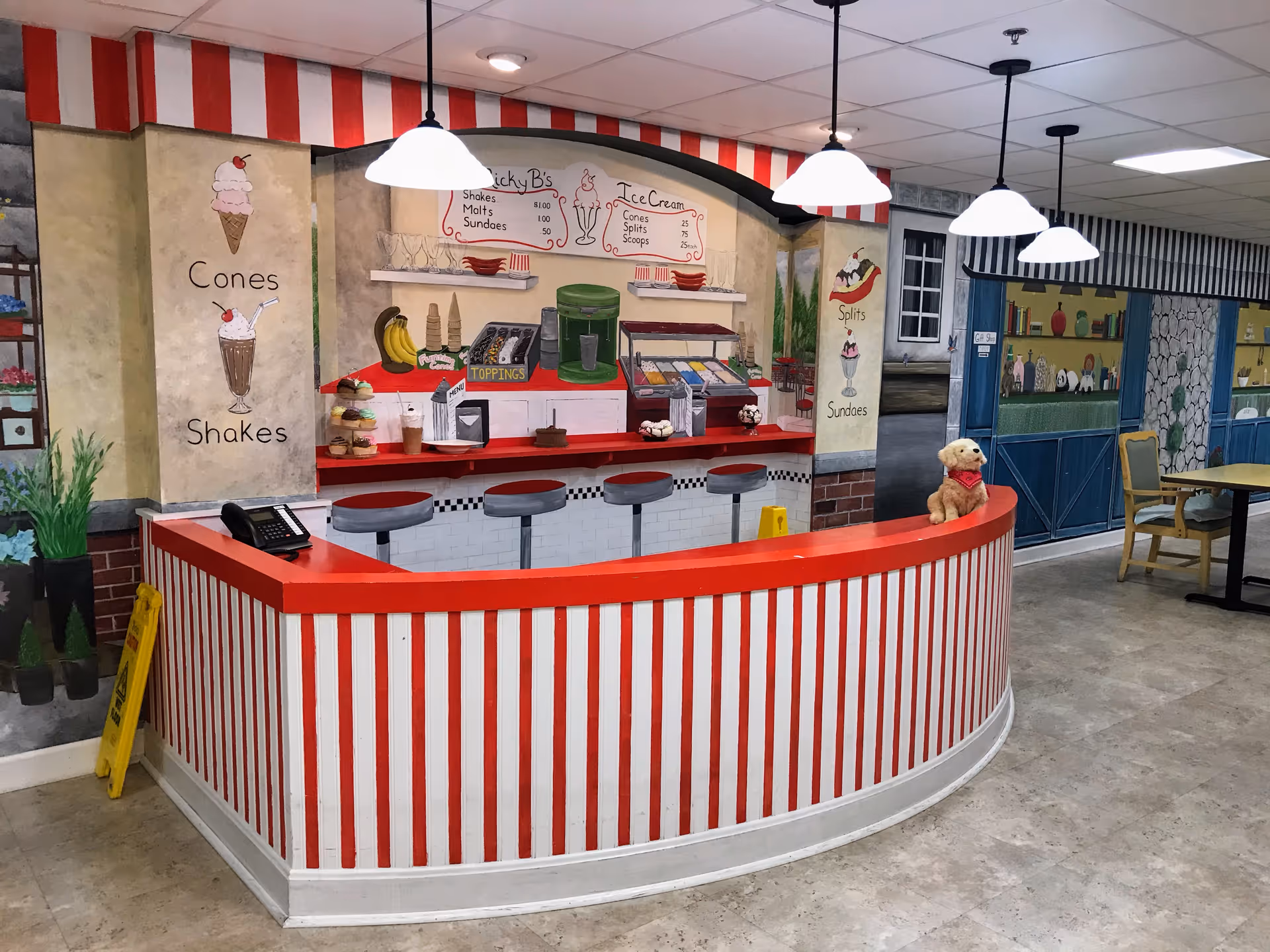 Interior of a senior living facility featuring a retro-style ice cream counter with red and white stripes, bar stools, and a mural depicting ice cream cones, shakes, sundaes, and toppings. There is a small stuffed dog on the counter and several hanging pendant lights above. In the background, there are tables and chairs with a mural of a storefront and garden scene on the walls.