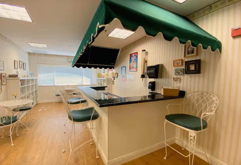 Interior view of a small dining or snack area with a counter and green cushioned bar stools. The counter has a black countertop and is topped with a green awning. The room has light-colored striped wallpaper and wooden flooring. There are small round tables with matching chairs, framed pictures on the walls, and a window with blinds letting in natural light.