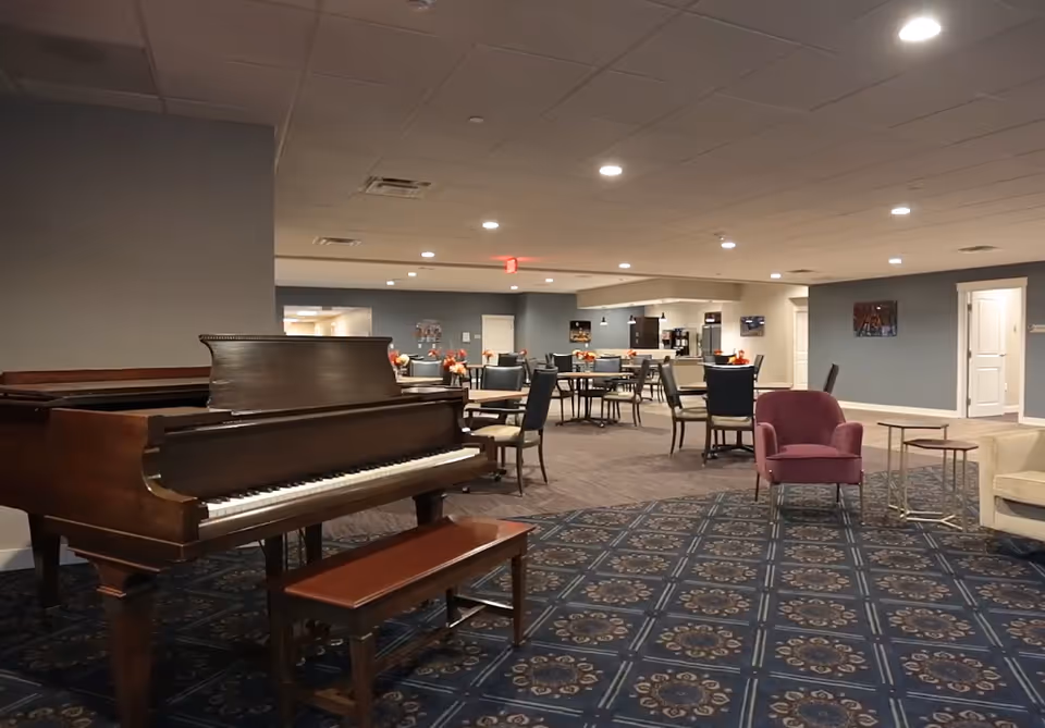 Interior view of a spacious common area in an assisted living facility featuring a grand piano with a bench on a patterned carpet, several round tables with chairs, a purple armchair, and a beige sofa. The room has a blue and gray color scheme with recessed lighting and a kitchenette area in the background.