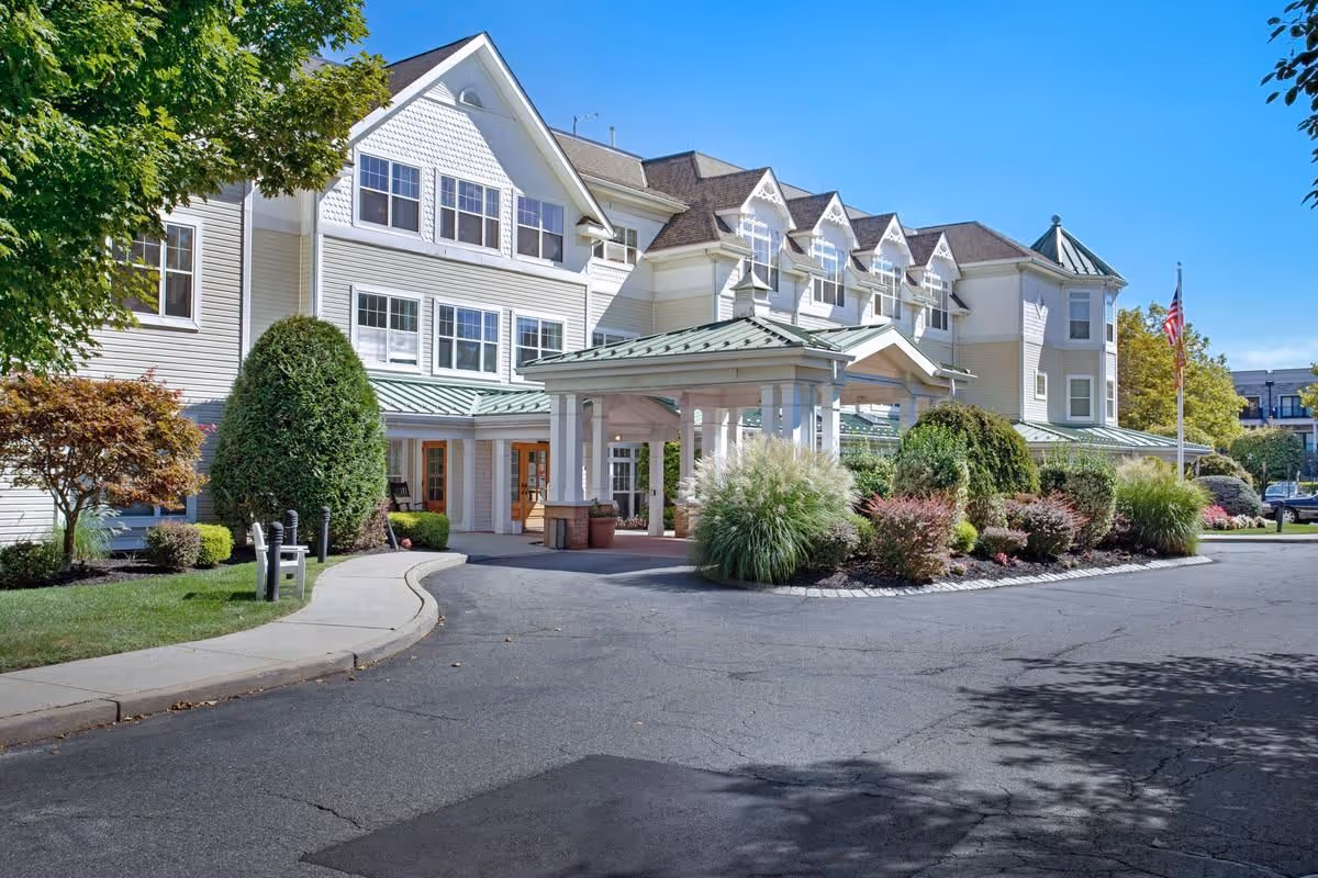 Front entrance of a multi-story senior living building with a covered porte-cochere, landscaped driveway, and an American flag.