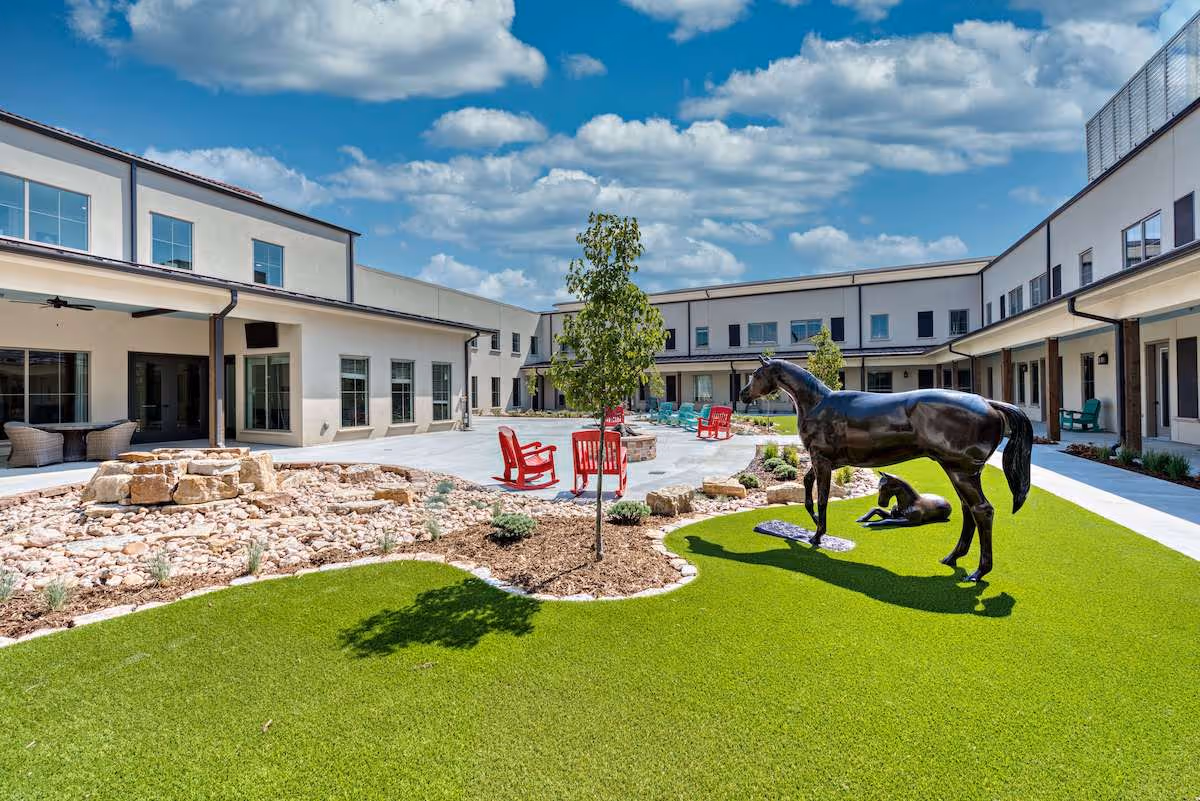 Outdoor courtyard area of a senior living facility with artificial grass, a small tree, red rocking chairs around a fire pit, and bronze statues of a horse and a foal. The courtyard is surrounded by a two-story building under a partly cloudy blue sky.