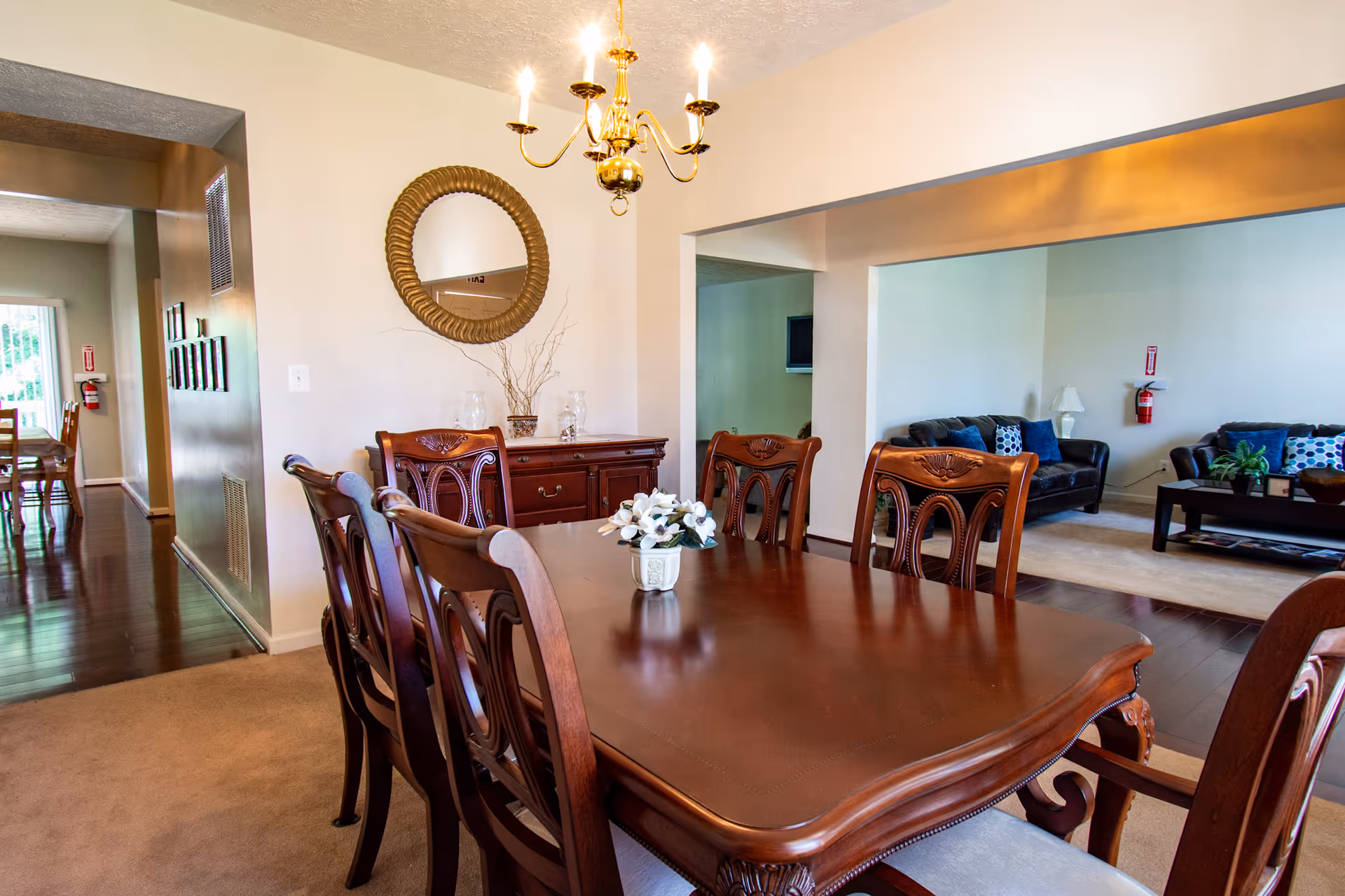 A dining room with a polished wooden table surrounded by six matching chairs. A small flower arrangement is placed in the center of the table. On the wall, there is a round decorative mirror above a wooden sideboard with glass vases and a plant. A brass chandelier with lit bulbs hangs above the table. In the background, an adjoining living room is visible with dark leather sofas, a coffee table, and a lamp. The floors are a mix of carpet and wood, and there is a hallway leading to another room.