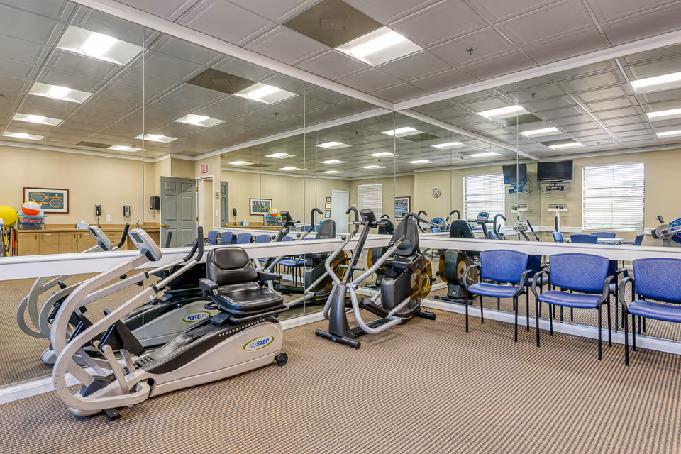 A fitness room with exercise equipment including a recumbent bike and an elliptical machine, blue chairs lined up against a mirrored wall, beige walls, and a carpeted floor. The room is well-lit with ceiling lights and has windows with blinds.
