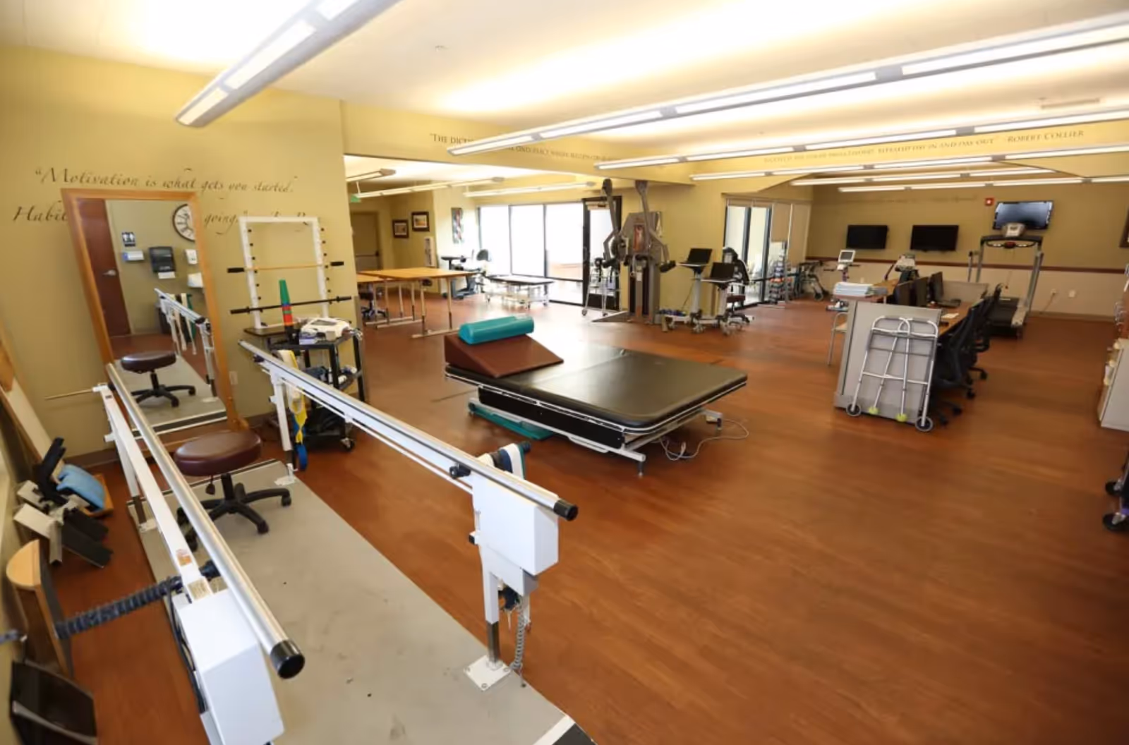 A spacious physical therapy room with wooden flooring, equipped with parallel bars for walking exercises, therapy tables, exercise equipment, chairs, and a walker. The room has large windows allowing natural light and motivational quotes on the walls.