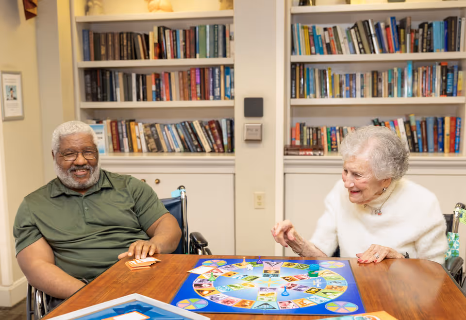 An elderly man and woman sitting in wheelchairs at a wooden table playing a colorful board game together in a room with bookshelves filled with books in the background. Both are smiling and appear to be enjoying the activity.
