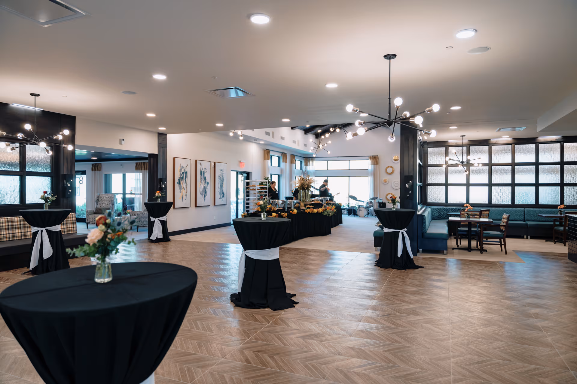 A spacious, well-lit event room with modern chandeliers and wooden flooring. Several tall cocktail tables covered with black tablecloths and white sashes are arranged around the room, each decorated with small floral arrangements. In the background, there is a buffet table with food and two people standing behind it. The room features large windows, framed artwork on the walls, and a seating area with tables and chairs along one side.