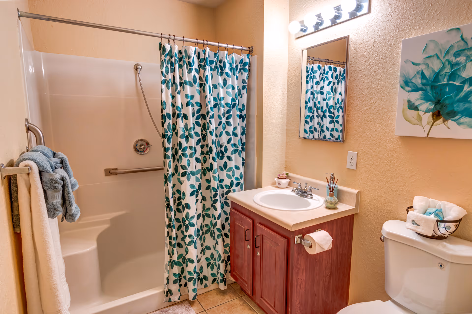Bathroom with a shower-tub behind a teal leaf-pattern shower curtain, a sink vanity with mirror, toilet, and towel rack.