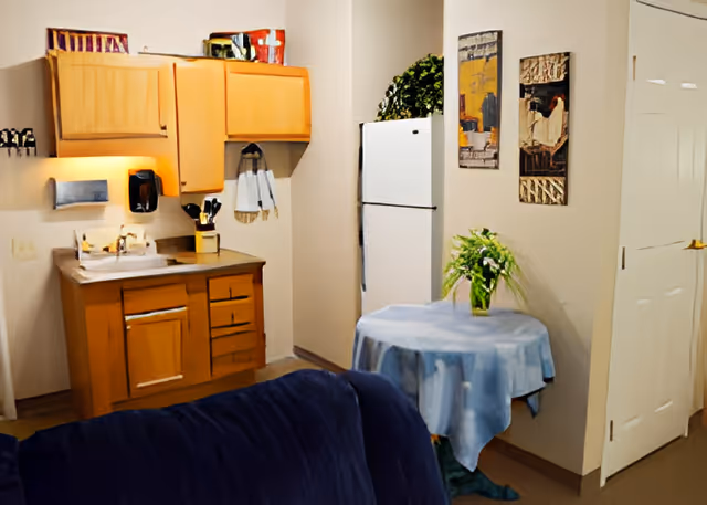 A small kitchen area with wooden cabinets, a countertop with a sink, a white refrigerator, and a round table covered with a blue tablecloth and a vase of flowers on top. There are two framed artworks on the wall and a dark blue couch partially visible in the foreground.
