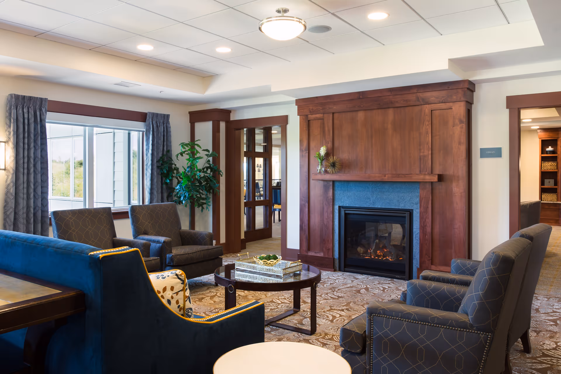 A cozy living room area in a senior living facility featuring a wooden fireplace with a small decorative plant on the mantel, surrounded by comfortable upholstered chairs and a blue sofa. There is a round coffee table in the center with decorative items, a large window with curtains, and a potted plant near the doorway leading to another room.