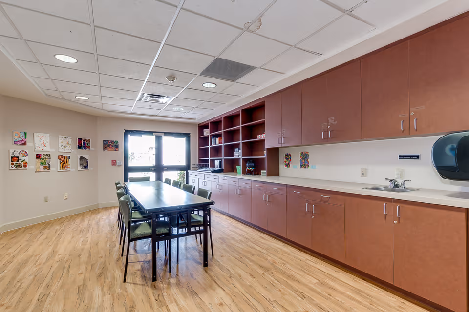 A bright room with a long table surrounded by chairs in the center. Along one wall, there are brown cabinets with a countertop, a sink, and open shelves holding various items. The floor is wood-patterned, and the ceiling has recessed lighting. Artwork is displayed on the opposite wall near a set of glass doors letting in natural light.