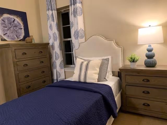 A bedroom with a single bed featuring a white upholstered headboard, blue quilted blanket, and decorative pillows. To the right of the bed is a wooden nightstand with a blue lamp and a small green plant. To the left is a tall wooden dresser with decorative items on top. A window with patterned curtains is visible behind the bed.