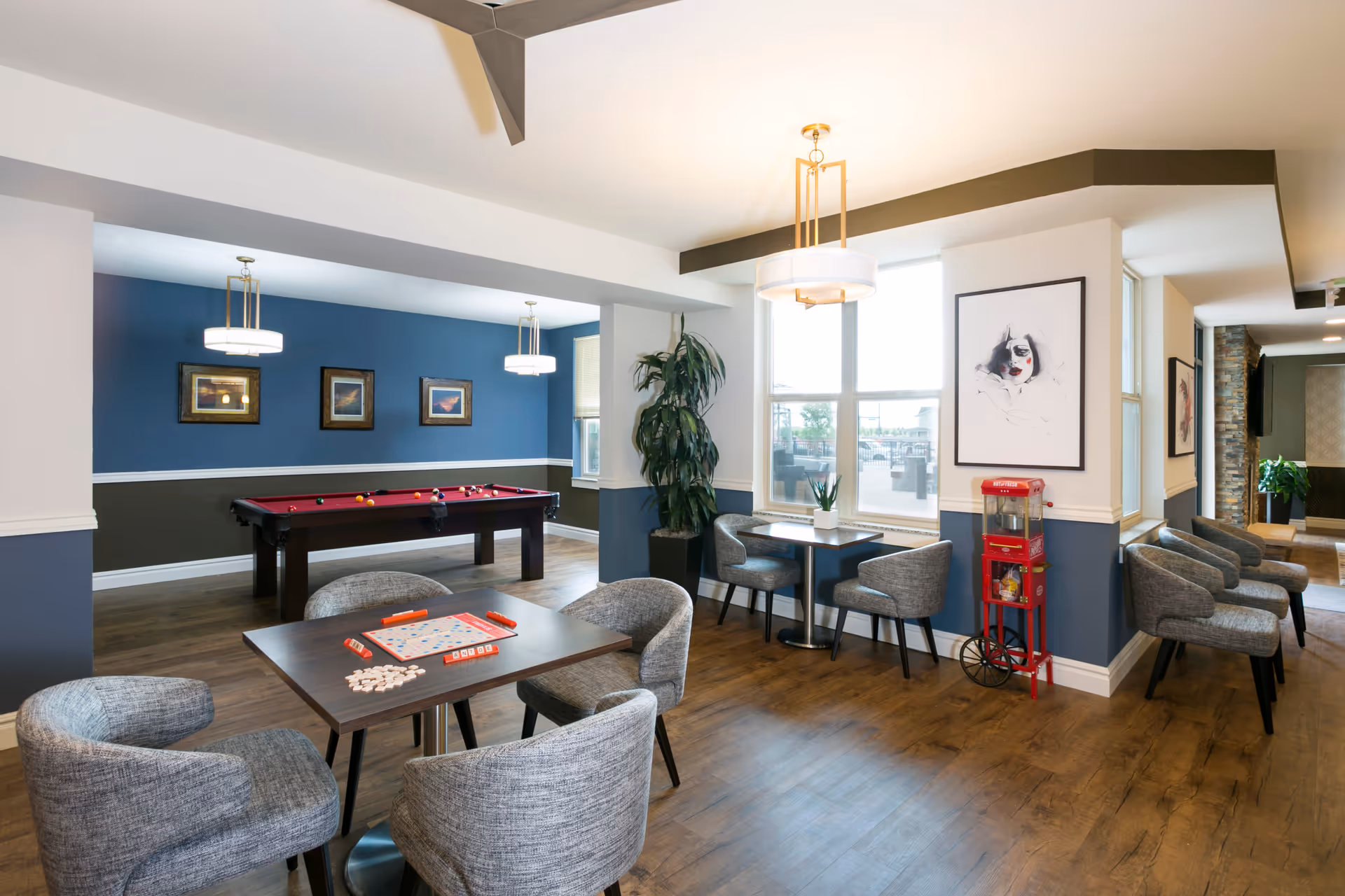 A bright and modern game room with a pool table in the background and two small tables with gray upholstered chairs. One table has a Scrabble game set up. The room features wood flooring, blue and white walls, large windows, contemporary light fixtures, framed artwork, a tall potted plant, and a red popcorn machine near the wall.
