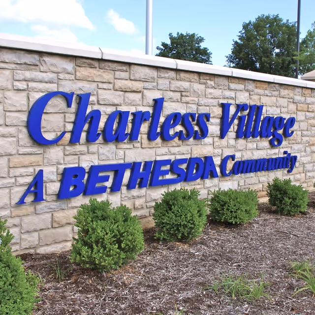 Stone entrance wall sign reading 'Charless Village A BETHESDA Community' with small shrubs and mulch in front.