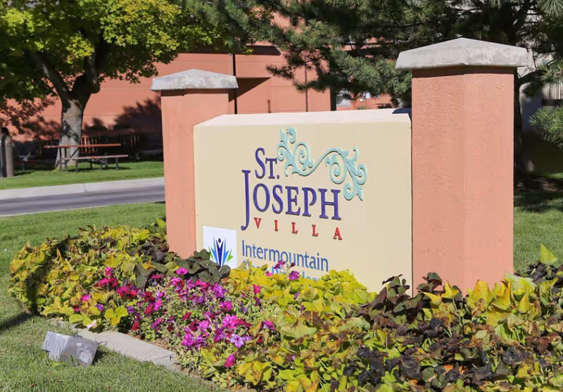 Outdoor view of a sign for St. Joseph Villa surrounded by green plants and purple flowers, with trees and a building in the background.