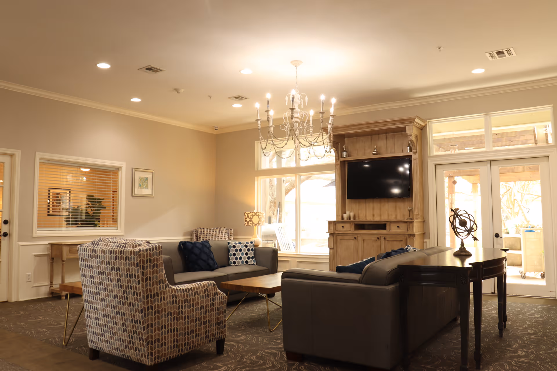 A cozy living room with patterned armchairs, a gray sofa with blue and white pillows, a wooden coffee table, and a wooden entertainment center with a mounted flat-screen TV. The room is softly lit by a chandelier and a table lamp, with large windows and glass doors letting in natural light.