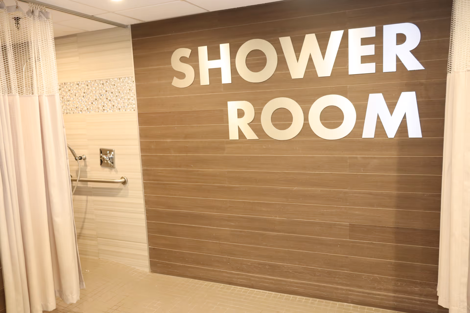 Interior view of a shower room with beige tiled walls and floor. There is a shower curtain on the left side and a metal grab bar on the wall. Large white letters on a wooden panel wall spell out 'SHOWER ROOM'.