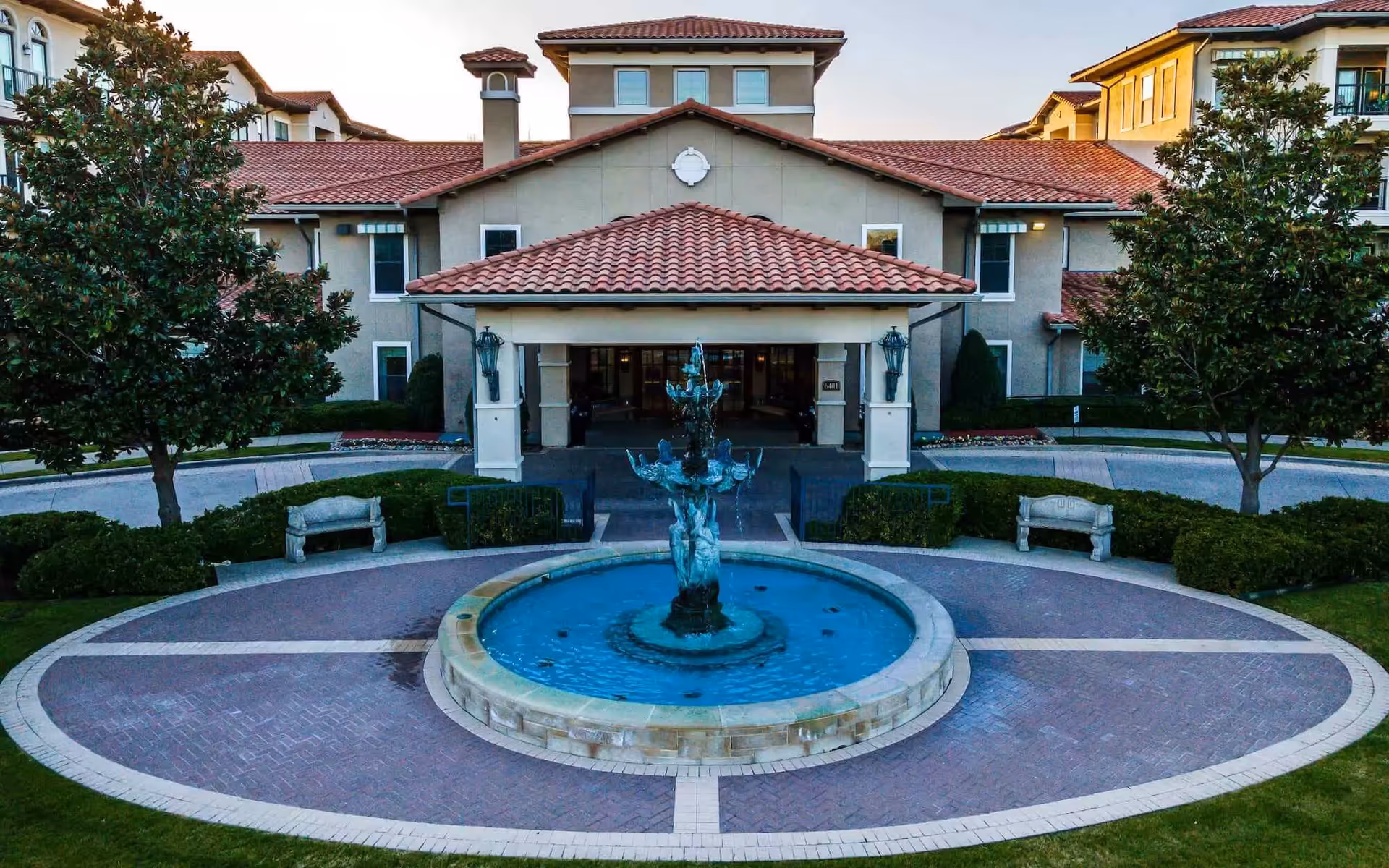 Front exterior view of Conservatory At Plano facility featuring a circular driveway with a central water fountain, two stone benches, manicured bushes, and trees on either side. The building has a beige facade with red tile roofing and multiple windows.