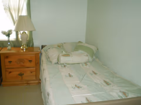 A small bedroom with a single bed covered in a light green and beige patterned comforter and matching pillows. Next to the bed is a wooden nightstand with two drawers, a white lamp with a beige shade, and a small vase with flowers. A window with light green curtains is partially visible behind the nightstand.