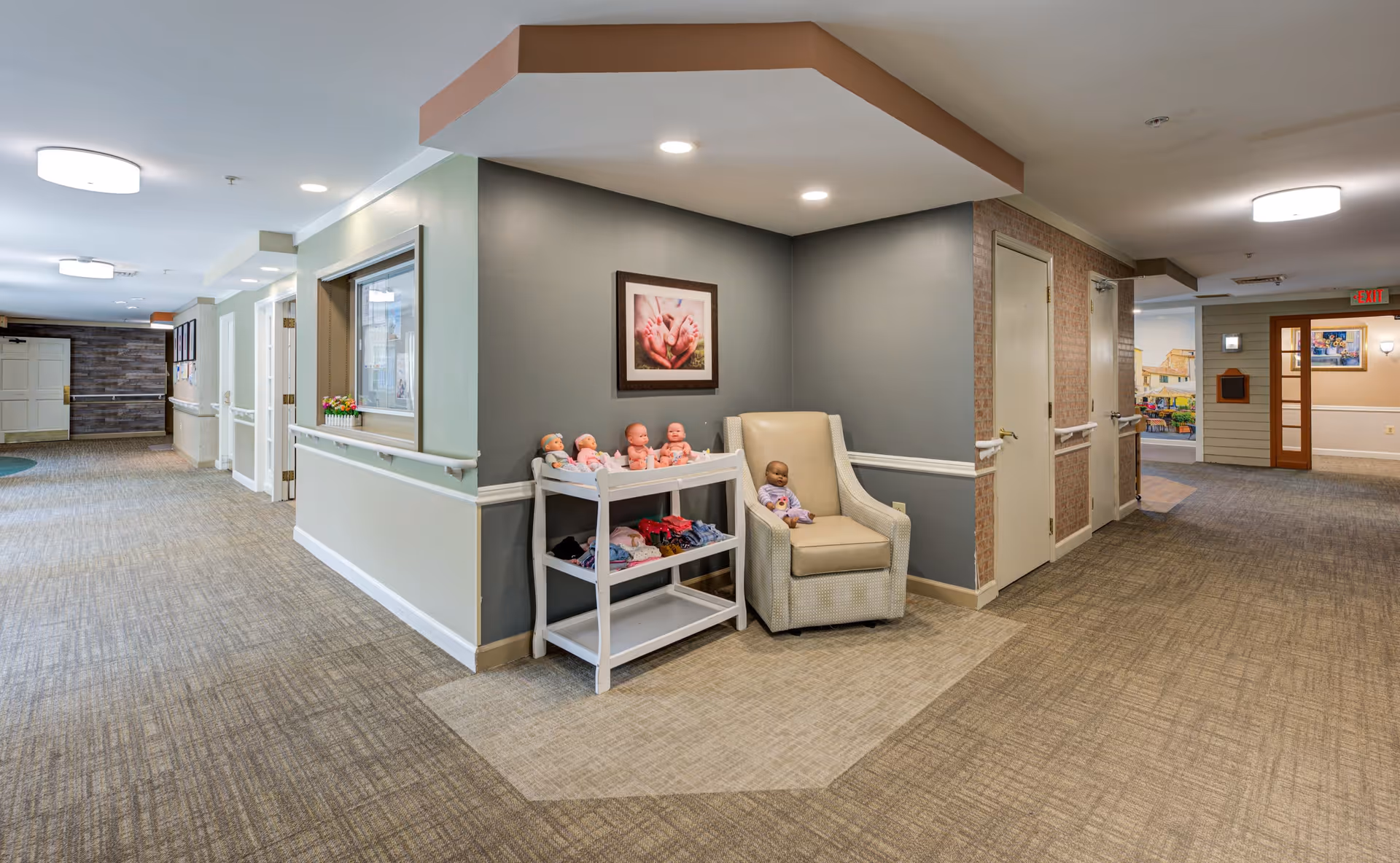 A well-lit hallway in a senior living facility with beige and gray walls and carpeted floors. In the corner, there is a beige armchair with a baby doll seated on it and a white shelf next to it holding several baby dolls and folded clothes. A framed picture of hands holding a baby is hung on the gray wall above the chair. The hallway has handrails along the walls and multiple doors leading to other rooms.