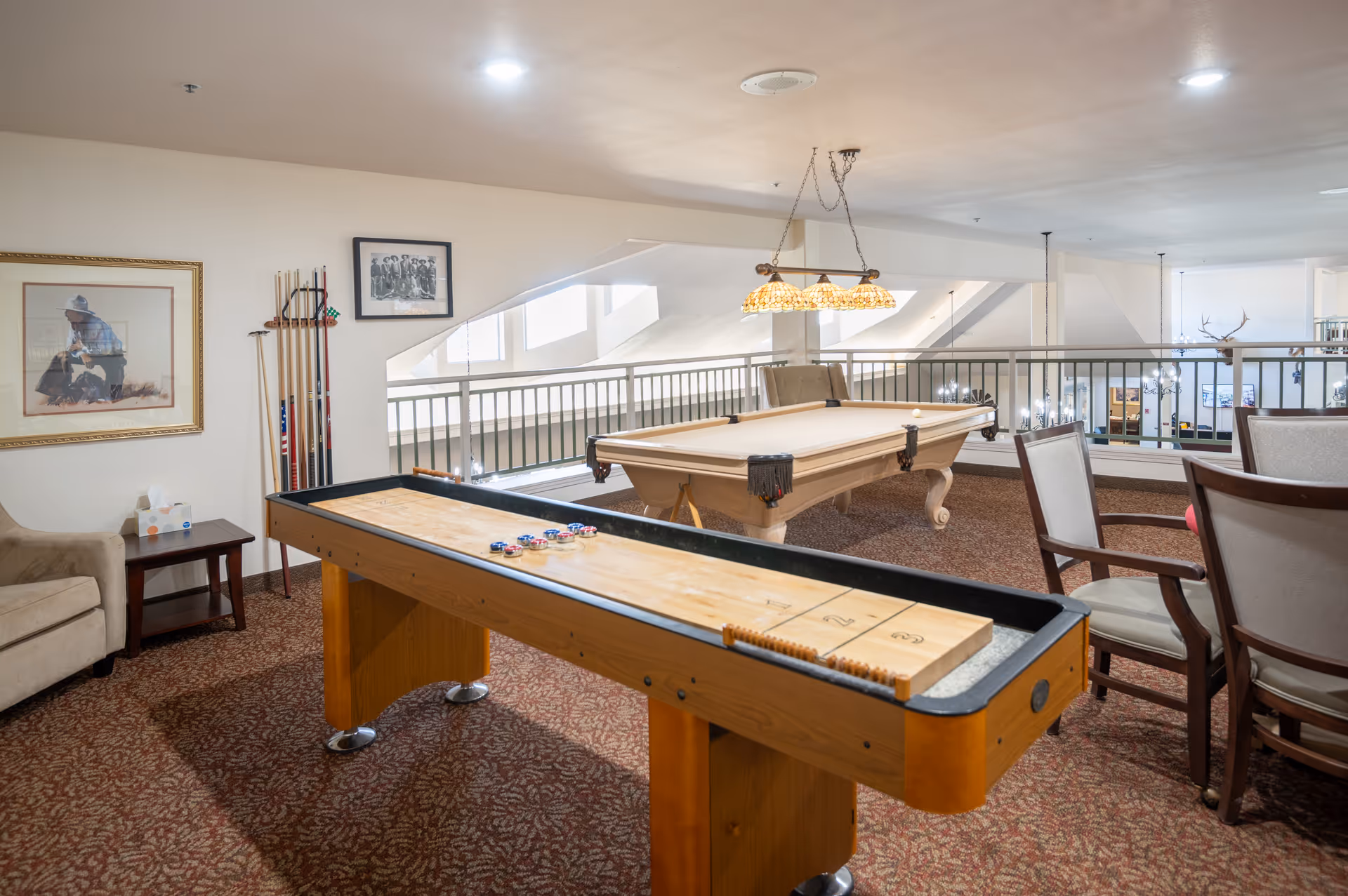 A recreational room in a senior living facility featuring a shuffleboard table in the foreground and a billiards table in the background. The room has carpeted flooring, a beige armchair, a small side table with a tissue box, framed artwork on the wall, and several chairs arranged near the tables. The space is well-lit with natural light coming through large windows and a hanging light fixture above the billiards table.