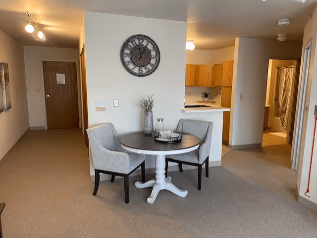 Interior view of a senior living apartment showing a small round dining table with two upholstered chairs. Behind the table is a wall with a large round clock. To the right is a kitchen area with wooden cabinets and a countertop. A hallway leads to a bathroom with a shower curtain visible. The floor is carpeted and the walls are painted light beige.