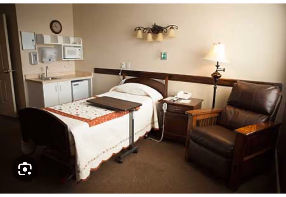 Single-occupancy senior bedroom with a hospital-style bed, overbed table, bedside nightstand, recliner chair and a small kitchenette area.
