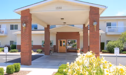 Entrance of a senior living facility named Sun Terrace Hermiston with a covered driveway supported by brick pillars, a building number 1550 displayed above, and yellow flowers in the foreground.