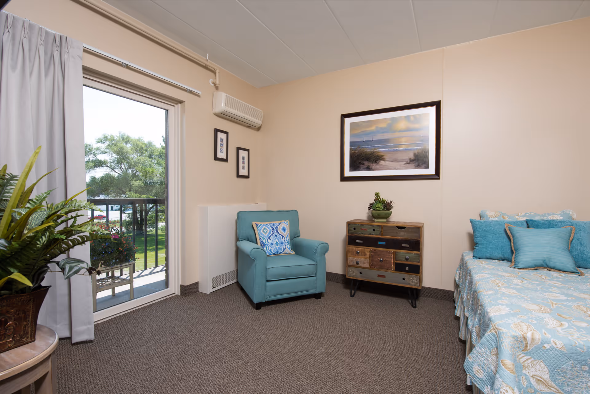 A cozy bedroom in an assisted living center featuring a bed with blue and beige seashell-patterned bedding and multiple pillows, a teal armchair with a decorative pillow, a small rustic wooden dresser with multiple drawers, a framed beach-themed painting on the wall, and a sliding glass door leading to a balcony with greenery outside.