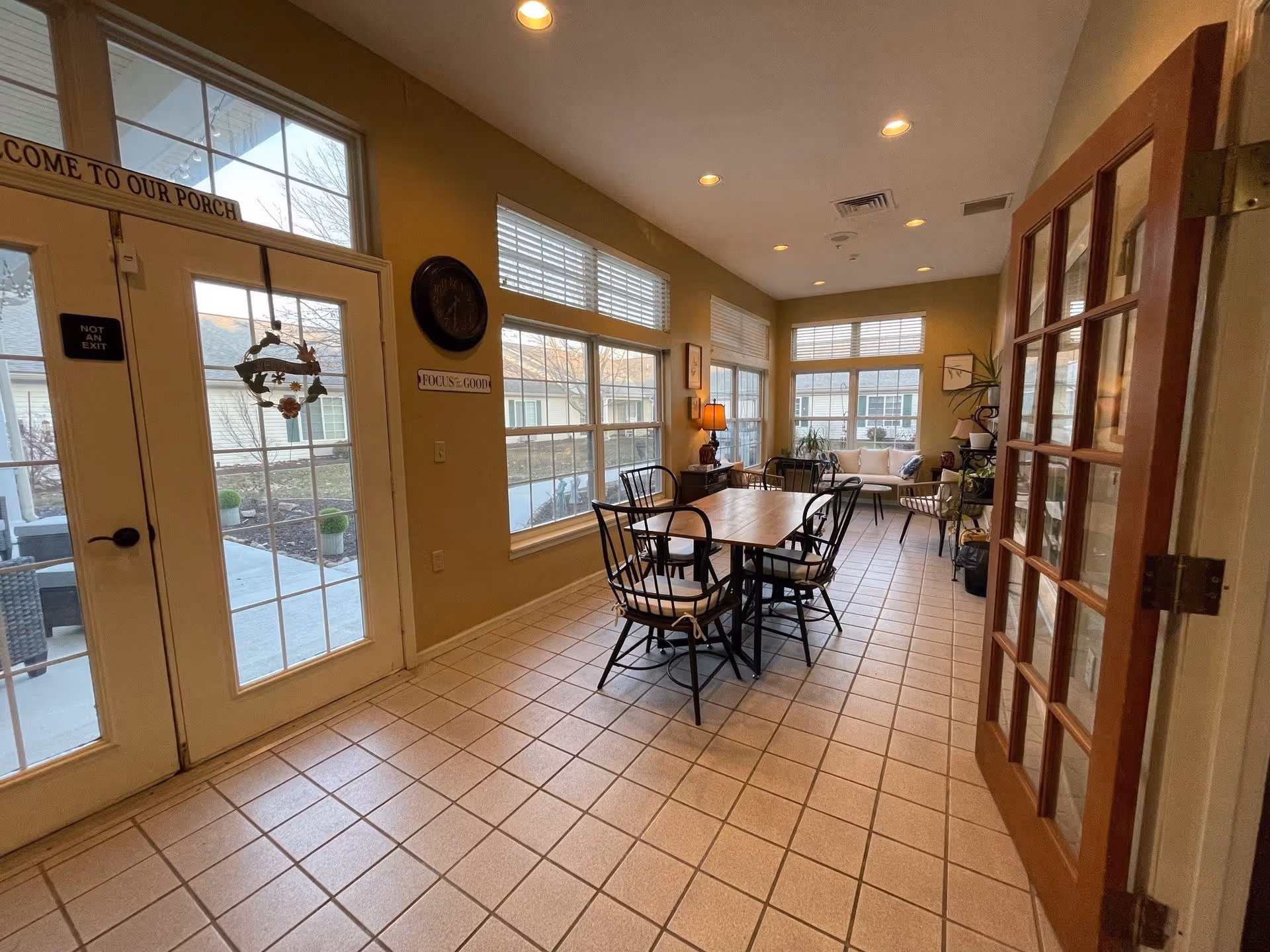 A bright sunroom with large windows on three sides, featuring a wooden table with six black metal chairs with cushions, a small sofa with pillows, a side table with a lamp, and various plants. The room has tiled flooring and recessed ceiling lights. A glass door with a sign that reads 'NOT AN EXIT' and a welcome wreath is visible on the left side.