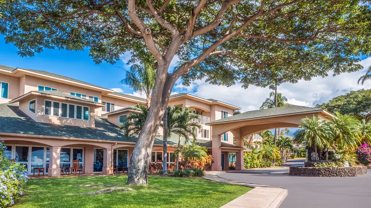 Exterior view of Holiday Kalama Heights facility showing a multi-story building with a covered entrance, surrounded by lush green trees, palm trees, and well-maintained landscaping under a bright blue sky.