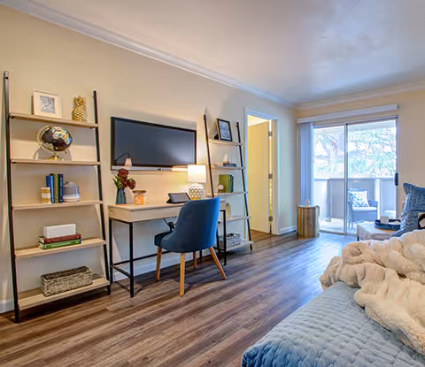 A cozy living room area with wooden flooring, featuring a blue cushioned chair at a desk with a lamp, bookshelves on either side of a wall-mounted flat screen TV, and a sliding glass door leading to a balcony with outdoor seating. A soft blanket is draped over a blue sofa in the foreground.