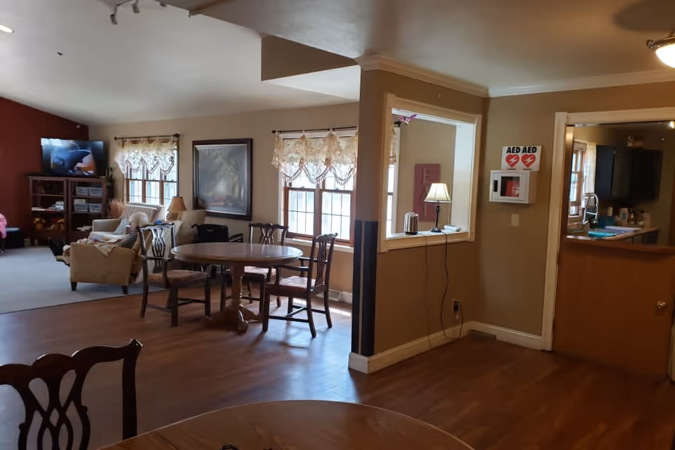 Interior view of a senior living facility showing a common area with wooden floors, a round dining table with chairs, a sitting area with armchairs and a TV, windows with lace curtains, a small lamp on a counter, and an AED device mounted on the wall near a kitchen entrance.