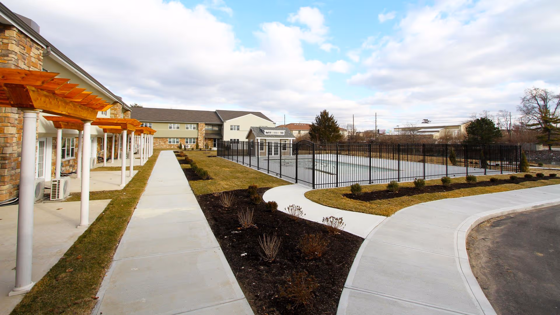 Landscaped courtyard with a paved walkway, wooden pergolas, and a fenced outdoor pool beside a senior living building under a partly cloudy sky.