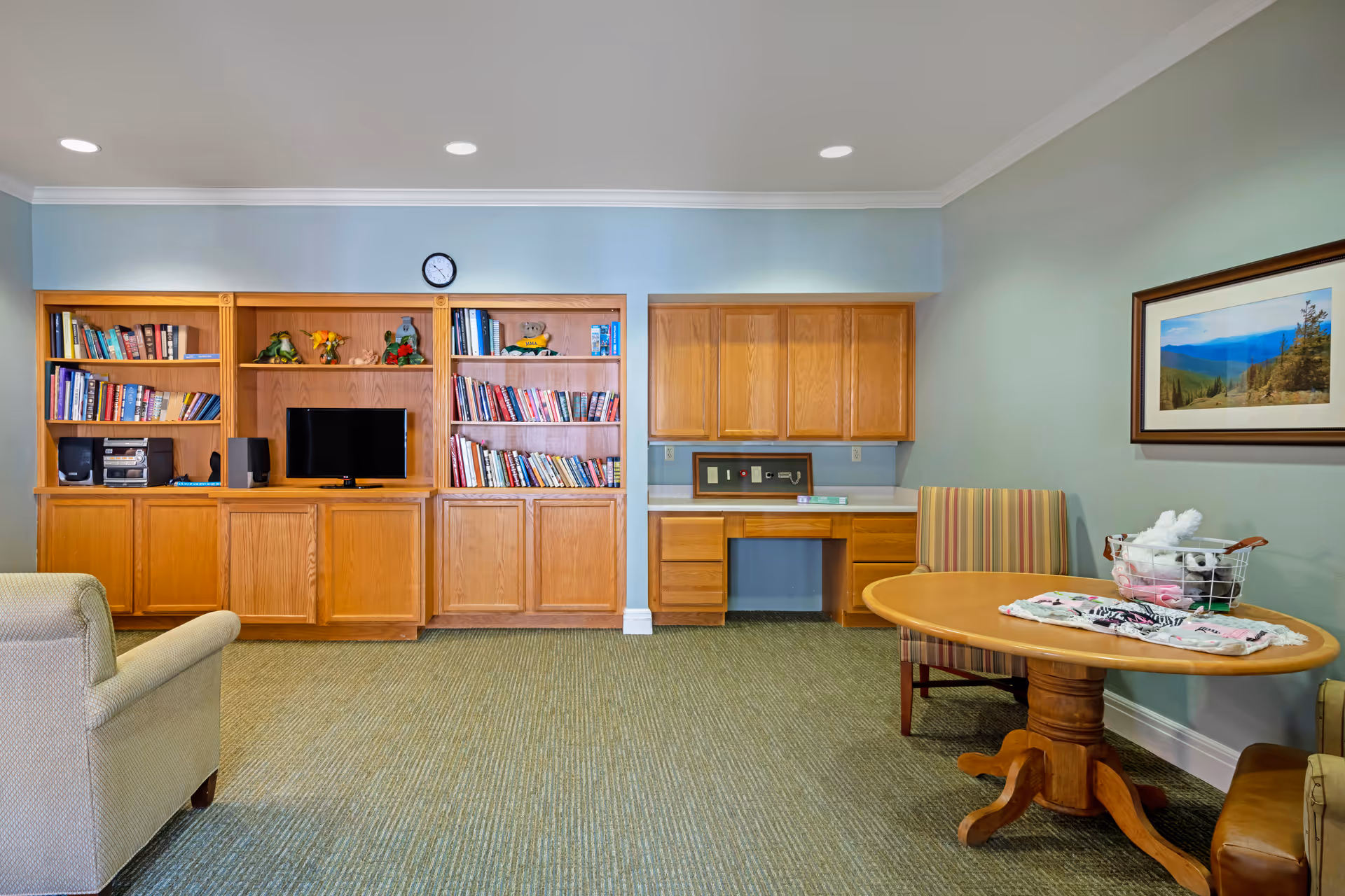 A cozy community room with built-in wooden bookshelves and cabinets, a TV, a desk, and a round table with chairs.
