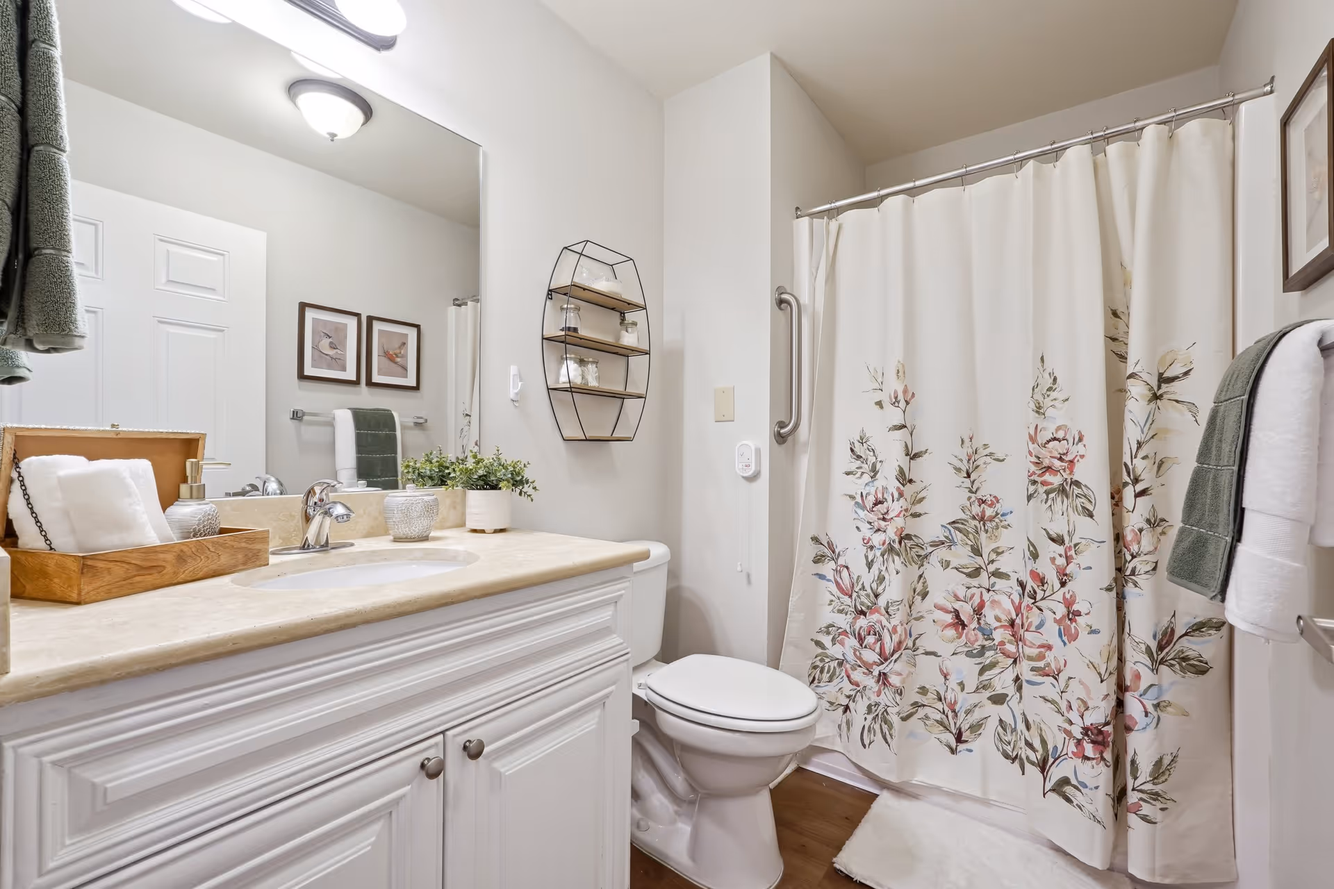 A clean and well-lit bathroom featuring a white vanity with a beige countertop, a sink with a silver faucet, and a wooden tray holding rolled white towels and a soap dispenser. Above the vanity is a large mirror with two light fixtures. To the right, there is a toilet next to a shower with a floral-patterned curtain. The walls are light-colored, with two framed pictures and a small wall-mounted shelf holding glass jars. Towels hang on racks near the shower.