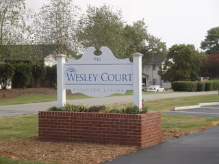 A white sign with blue text reading 'Wesley Court Assisted Living' mounted on two white posts above a red brick base, located near a driveway with trees and a building in the background.