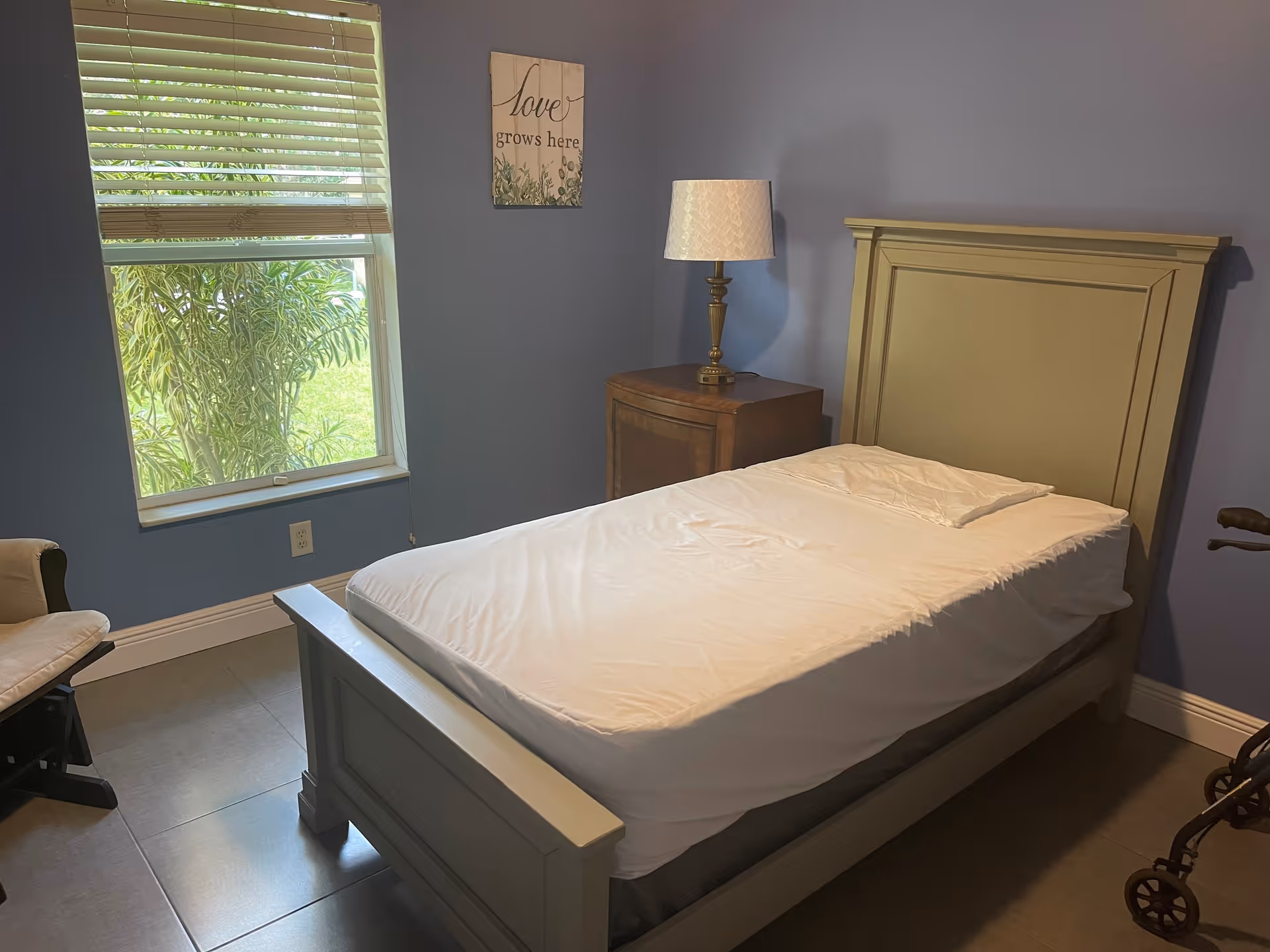A small bedroom with a single bed made with white sheets, a bedside table and lamp, a window with blinds, and a walker near the foot of the bed.