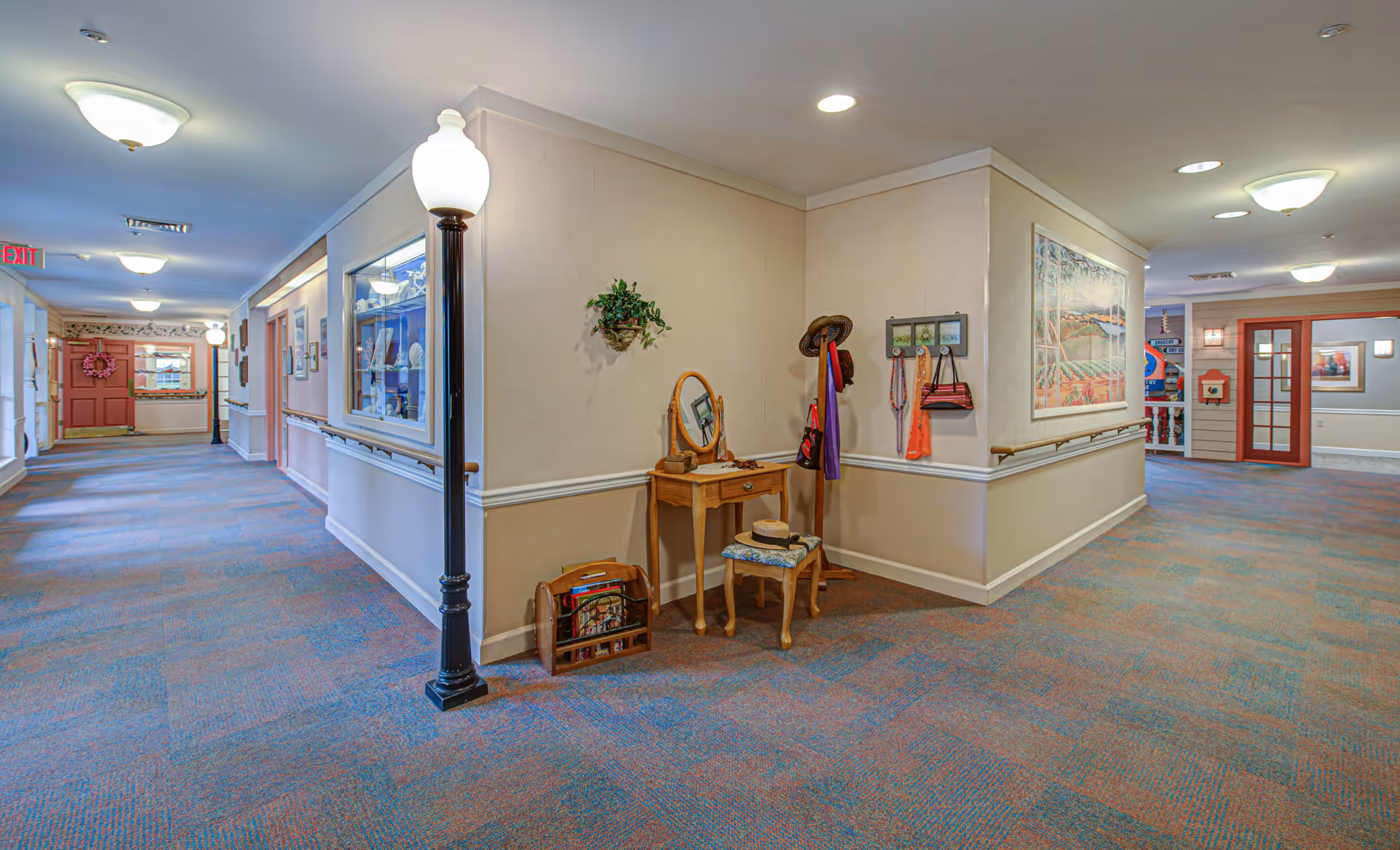 Interior hallway of a senior living facility with carpeted floors, beige walls, and ceiling lights. There is a small wooden vanity table with a mirror, a stool, a coat rack with hats and bags, and a magazine rack. The hallway has handrails along the walls and decorative streetlamp-style lights. Artwork and framed pictures are displayed on the walls.