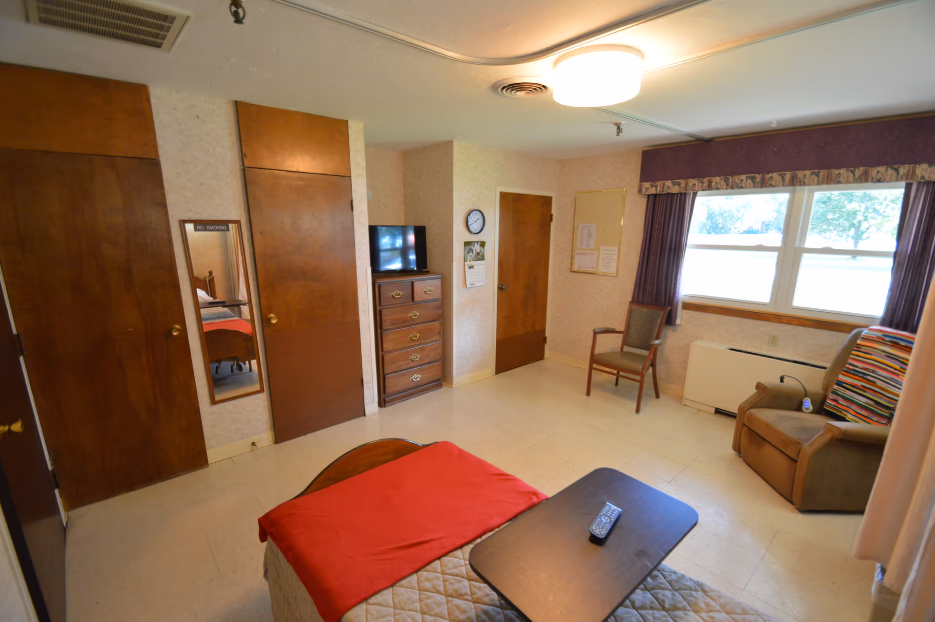 A simple bedroom in a senior living facility with a bed covered in a beige quilt and red blanket, a small dark wooden table with a remote control on it, a wooden dresser with a TV on top, a chair near a window with purple curtains, and two wooden doors on the left wall. A mirror with a 'No Smoking' sign is mounted between the doors.