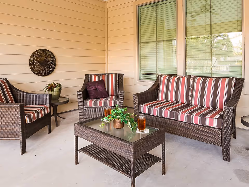 Covered patio with wicker seating and striped cushions, a glass-top coffee table holding drinks and a potted plant.